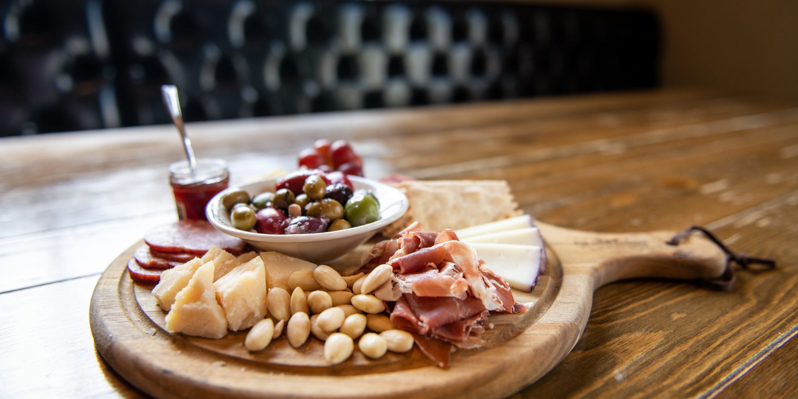 Wooden charcuterie board with nuts, cheeses, meats, crackers, olives, and jam.