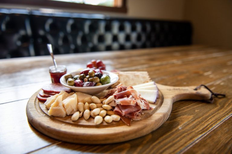 Wooden charcuterie board with nuts, cheeses, meats, crackers, olives, and jam.